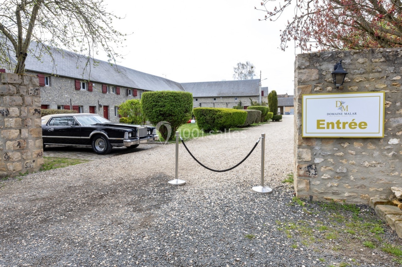 Entrée d'un domaine avec une allée gravillonnée, une voiture ancienne noire et un panneau indiquant ’Entrée’.
