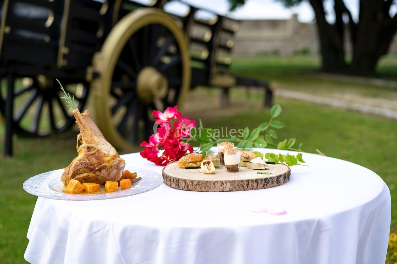 Assiette de côtelettes d'agneau et plateau de bouchées sur une table blanche en extérieur, près d'une charrette ancienne.