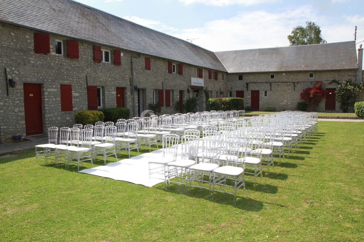 Chaises transparentes alignées sur une pelouse devant des bâtiments en pierre avec volets rouges, sous un ciel dégagé.