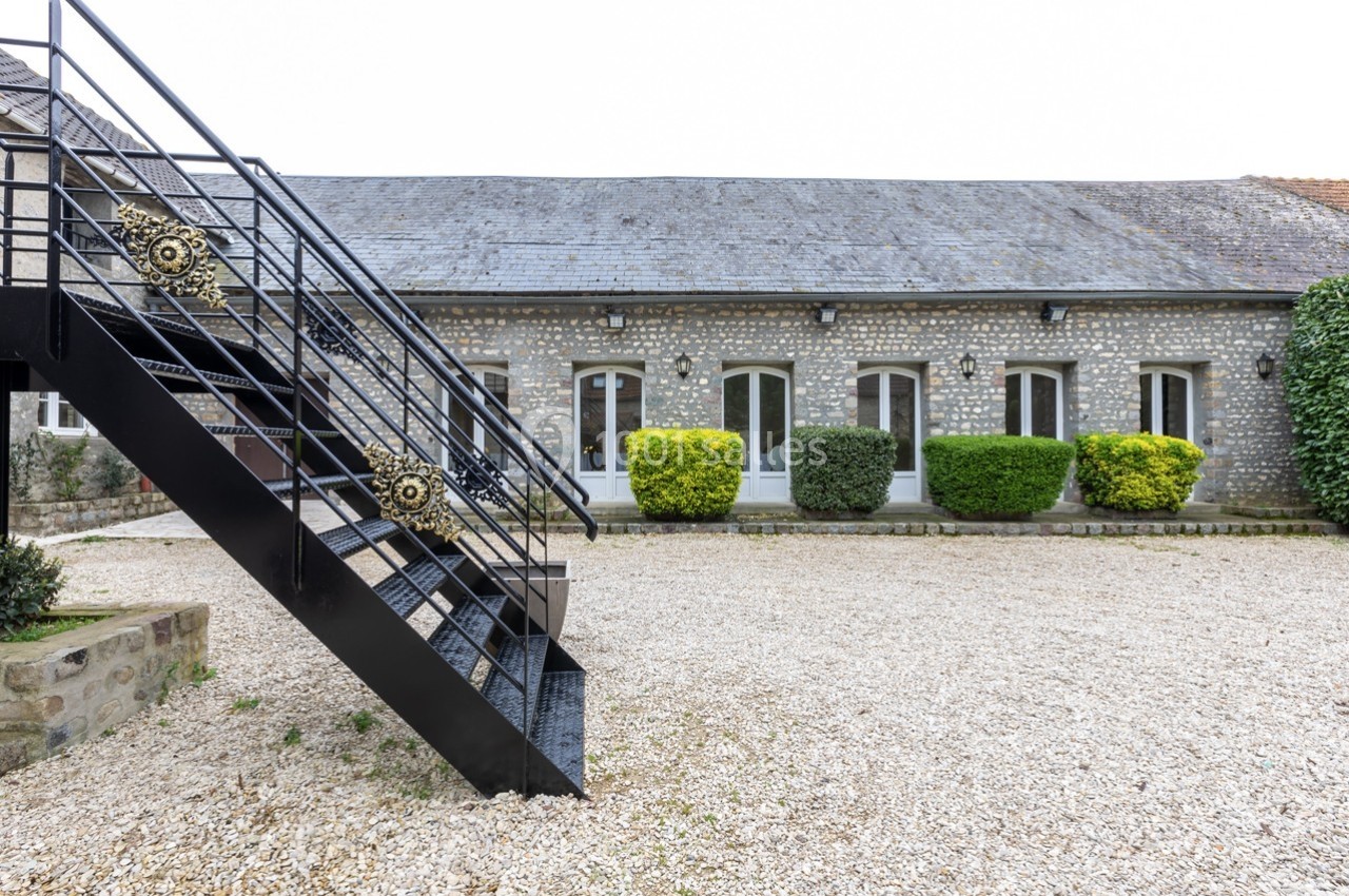 Façade en pierre d'un bâtiment avec des fenêtres cintrées, escalier métallique noir et cour gravillonnée.