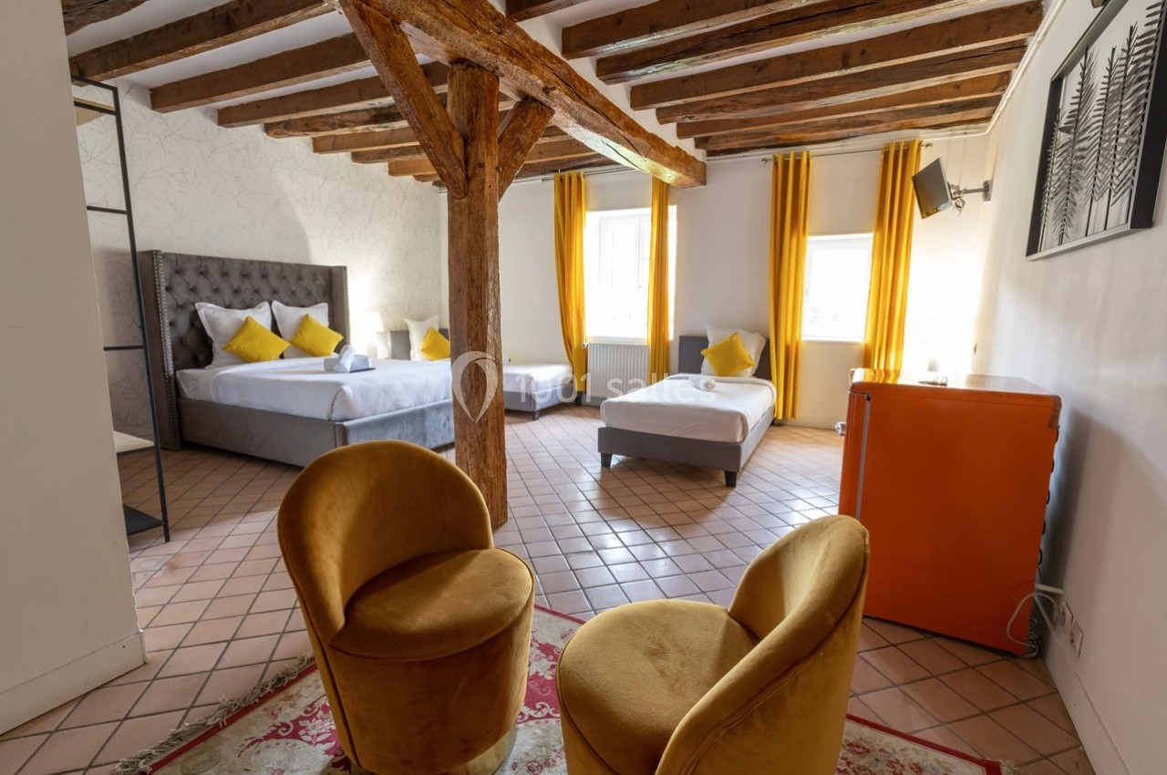 Chambre lumineuse avec poutres apparentes, deux lits, fauteuils jaunes, rideaux assortis et un mini-réfrigérateur orange.