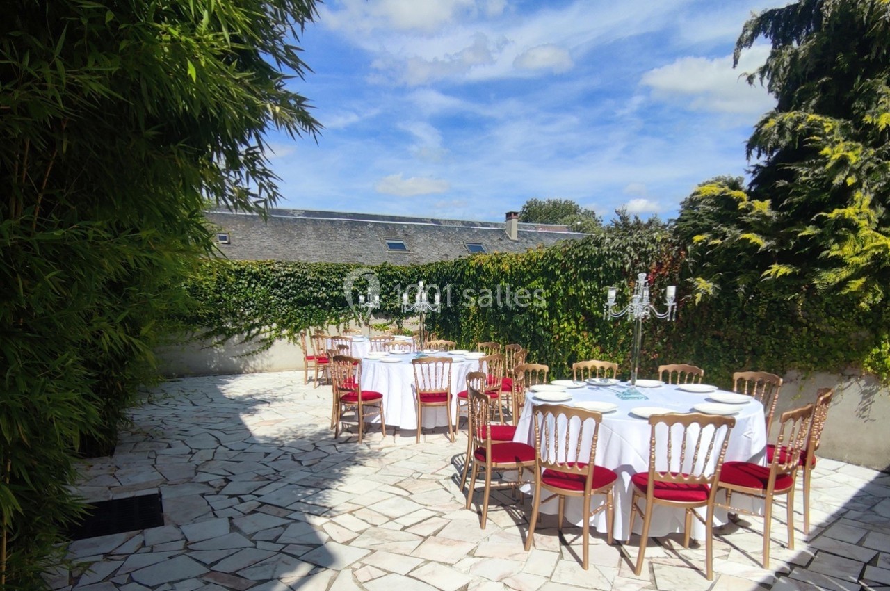 Tables rondes dressées avec nappes blanches et chaises rouges sur une terrasse pavée entourée de verdure.