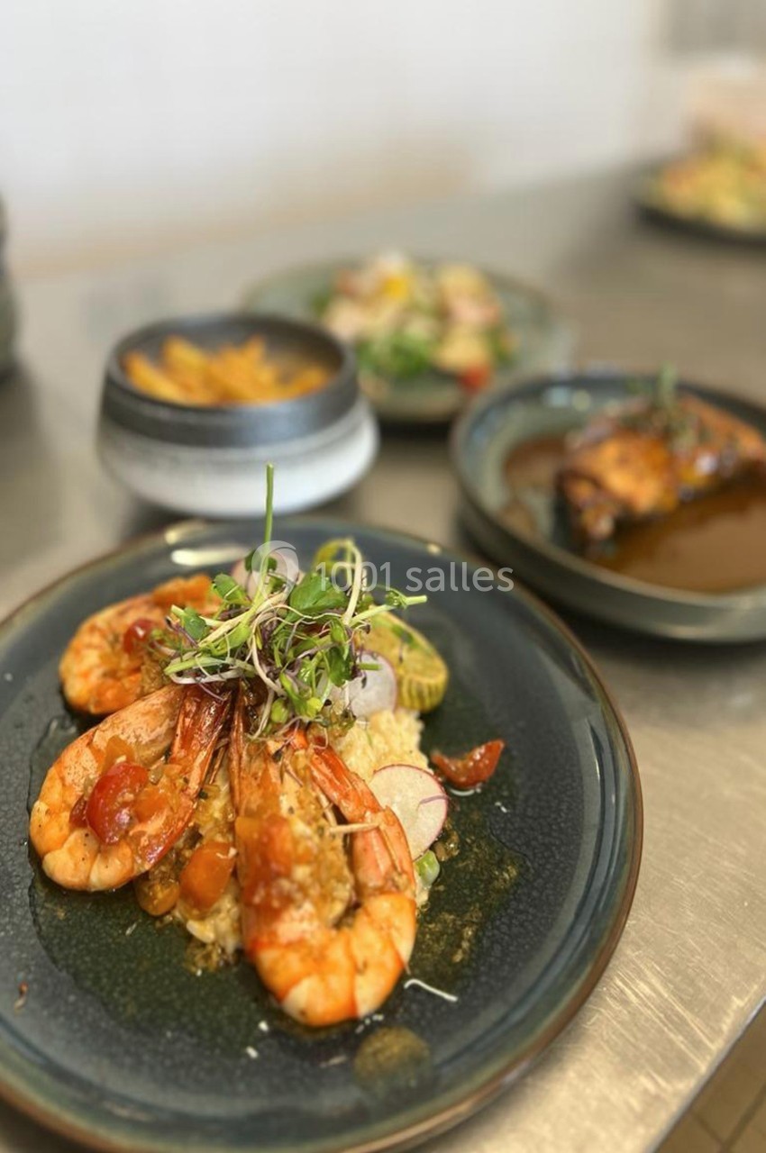 Assiette de crevettes grillées servies avec légumes et herbes fraîches, accompagnée de plats en arrière-plan.