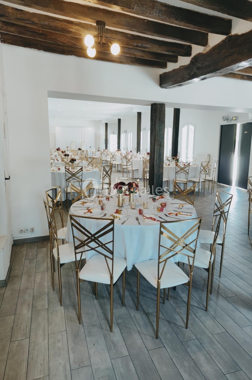 Salle de réception lumineuse avec tables rondes dressées, chaises dorées et poutres apparentes au plafond.