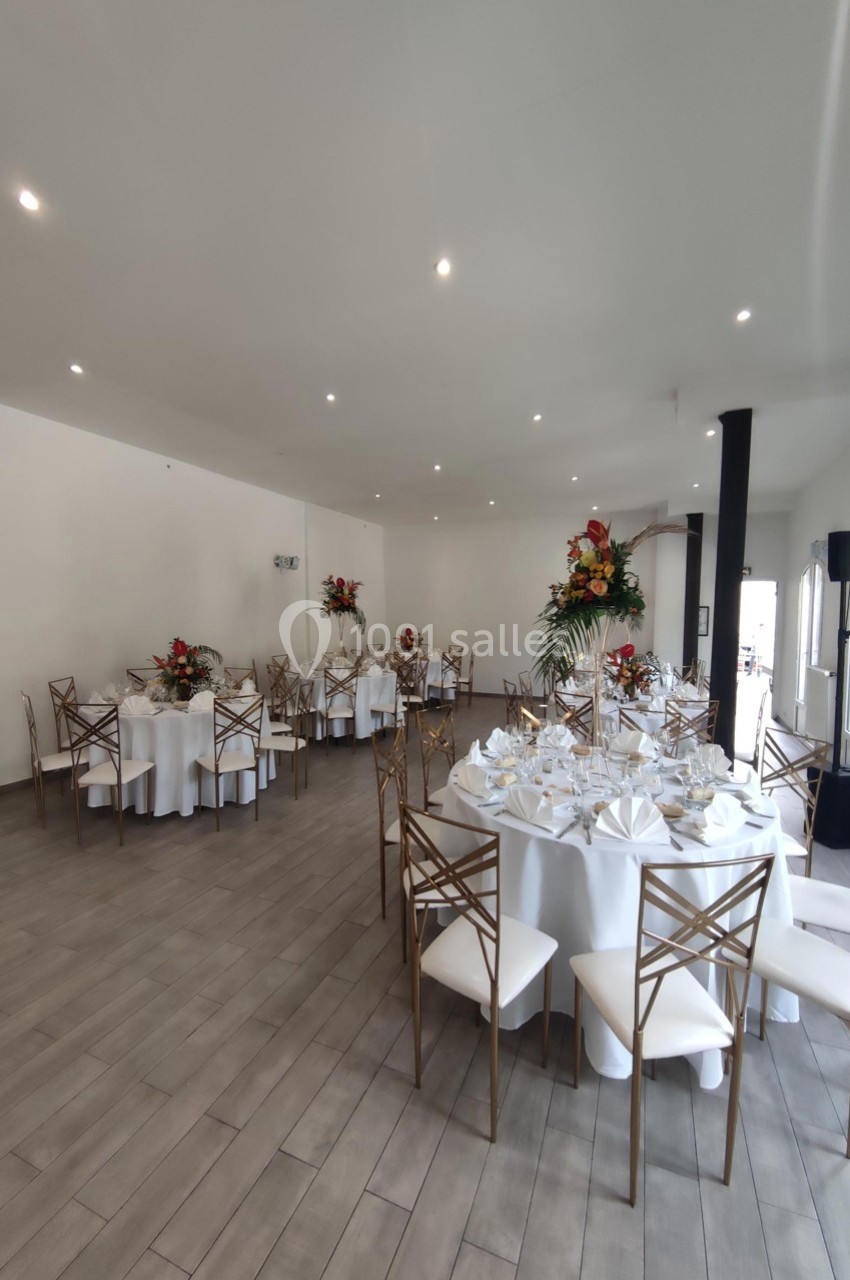 Salle de réception lumineuse avec tables rondes dressées, nappes blanches et décorations florales colorées.