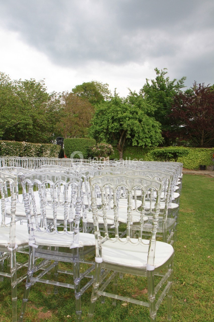 Chaises transparentes alignées sur une pelouse, entourées de haies et d'arbres sous un ciel nuageux.