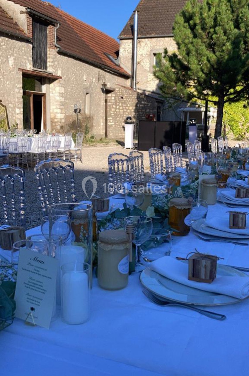 Tables dressées pour un mariage en extérieur, avec nappes blanches, bougies et décorations, devant des bâtiments en pierre.