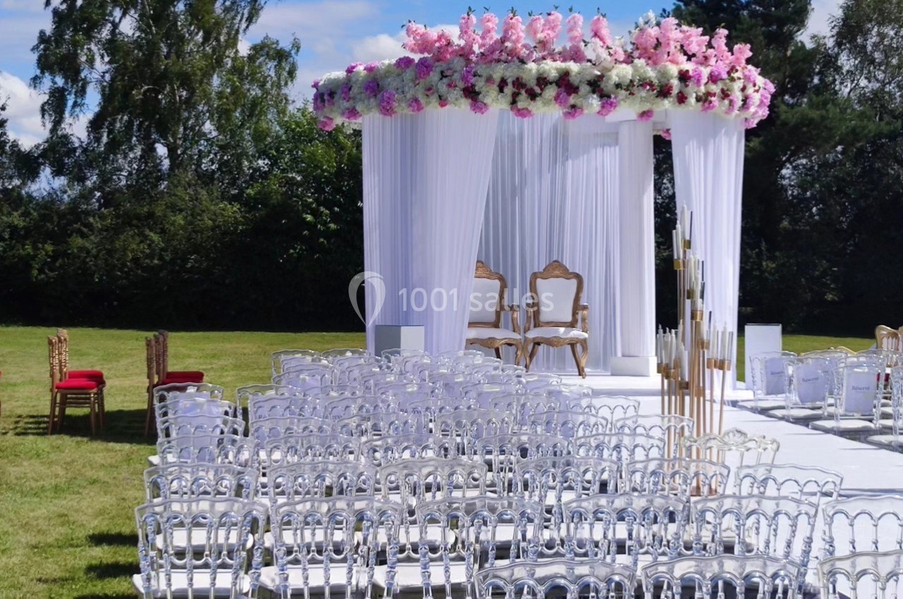Structure de cérémonie en plein air avec arche décorée de fleurs roses et blanches, entourée de chaises transparentes.