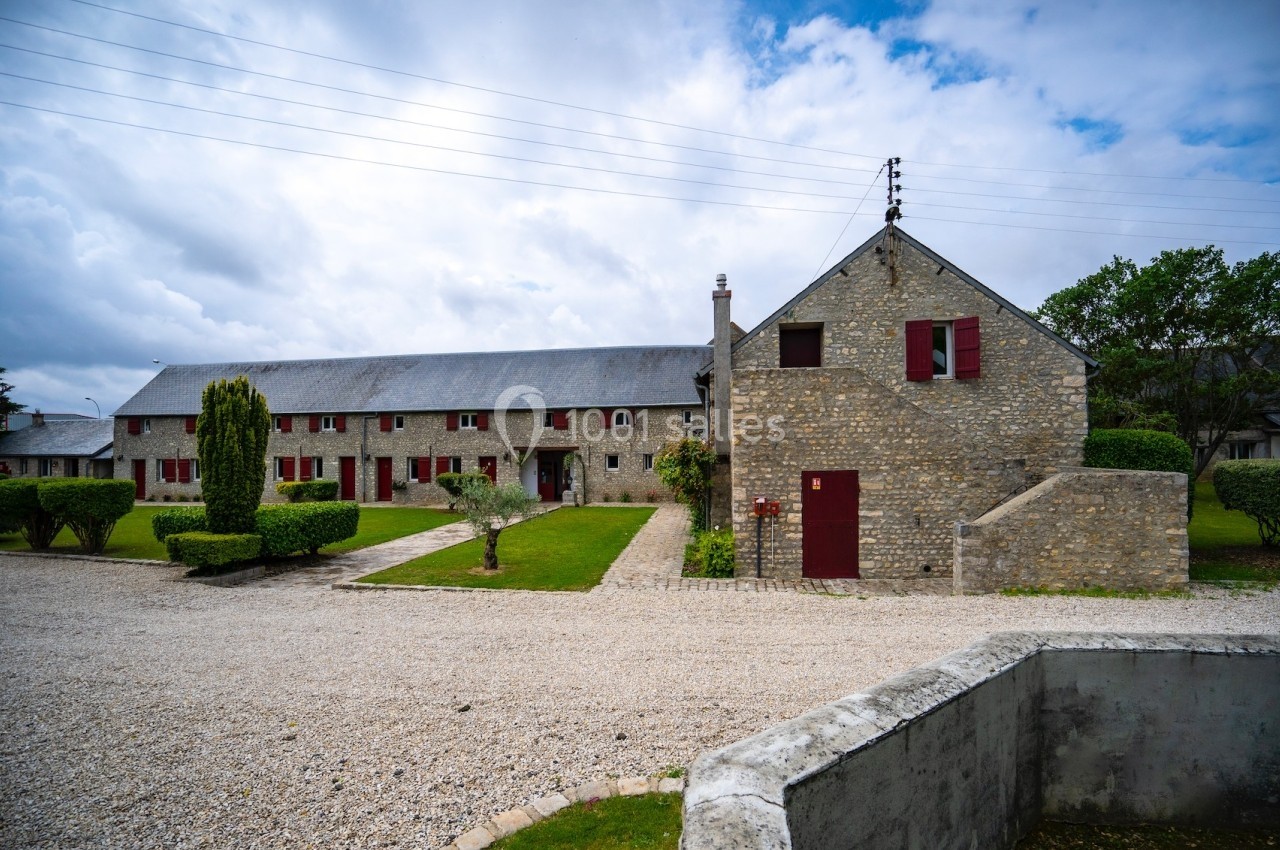 Bâtiment en pierre avec volets rouges, entouré d'une cour gravillonnée et d'un jardin aménagé sous un ciel nuageux.