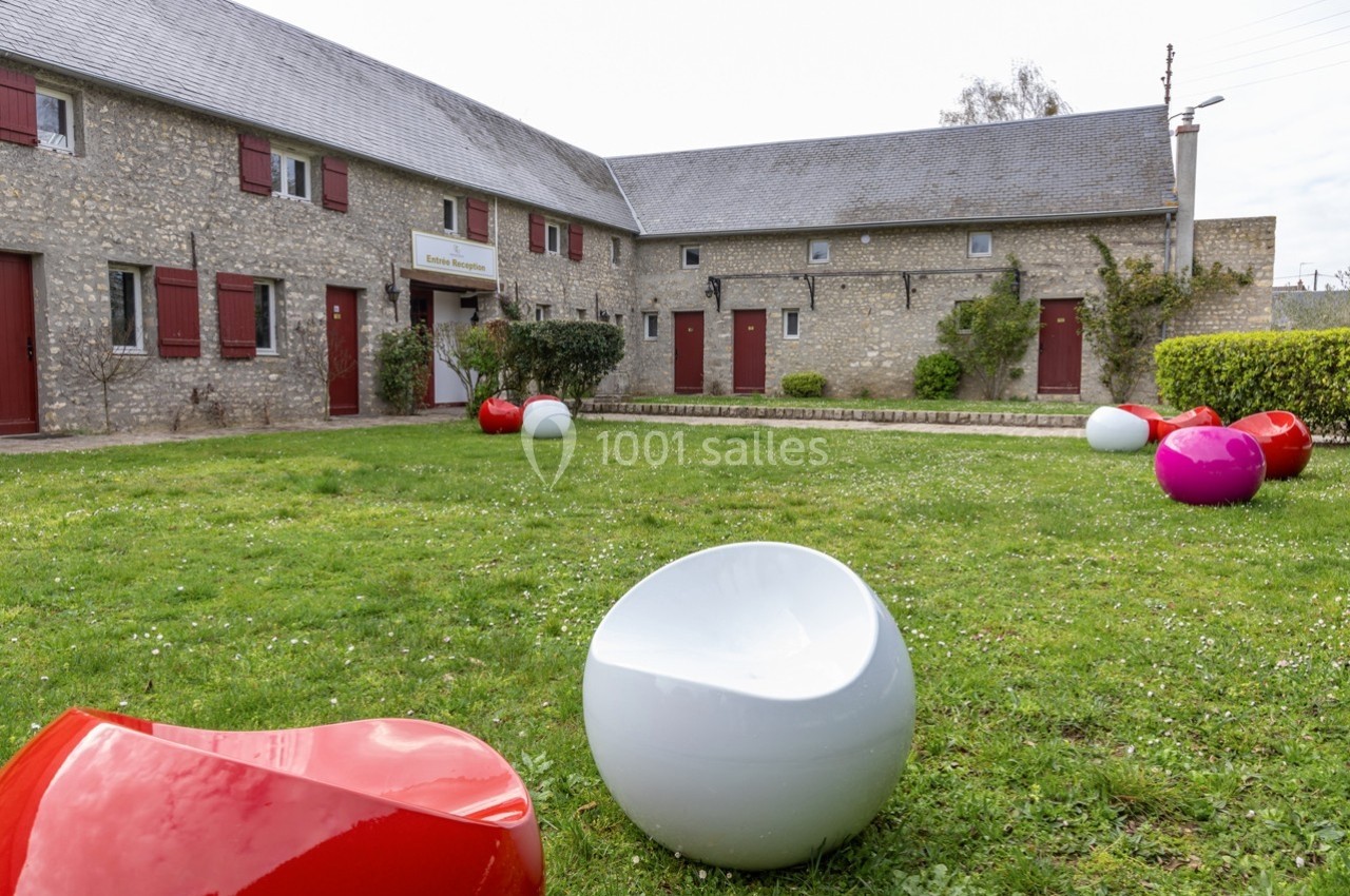 Cour enherbée avec des bâtiments en pierre, des volets rouges et des sièges colorés disposés sur la pelouse.