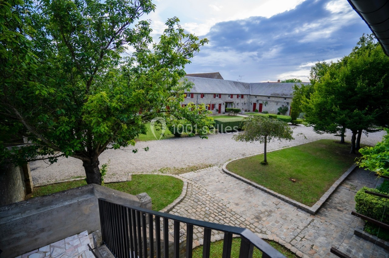 Cour pavée entourée de jardins et d'un bâtiment en pierre avec des volets rouges, vue depuis un escalier.