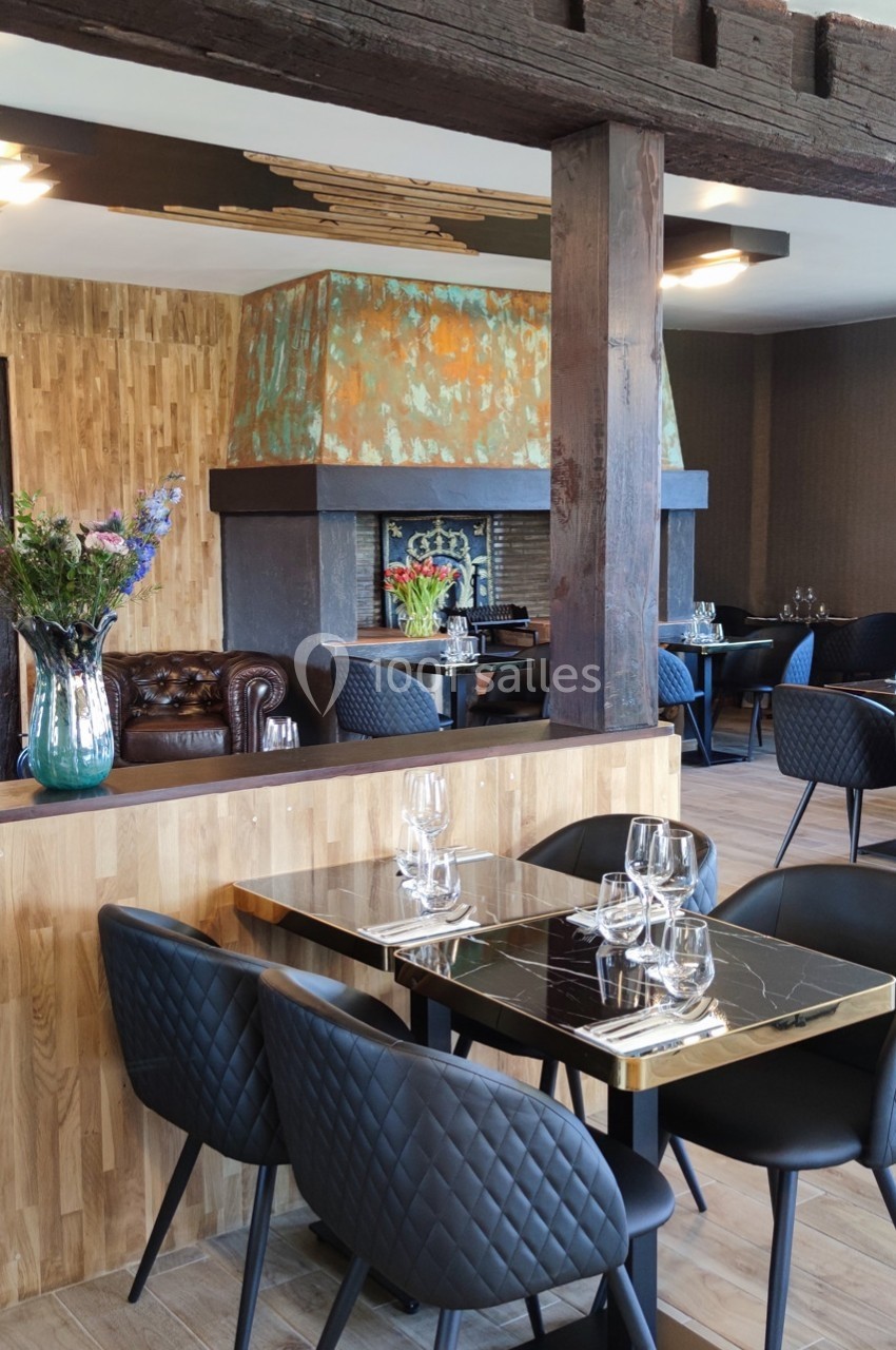 Salle de restaurant moderne avec tables dressées, chaises noires matelassées et cheminée en arrière-plan.