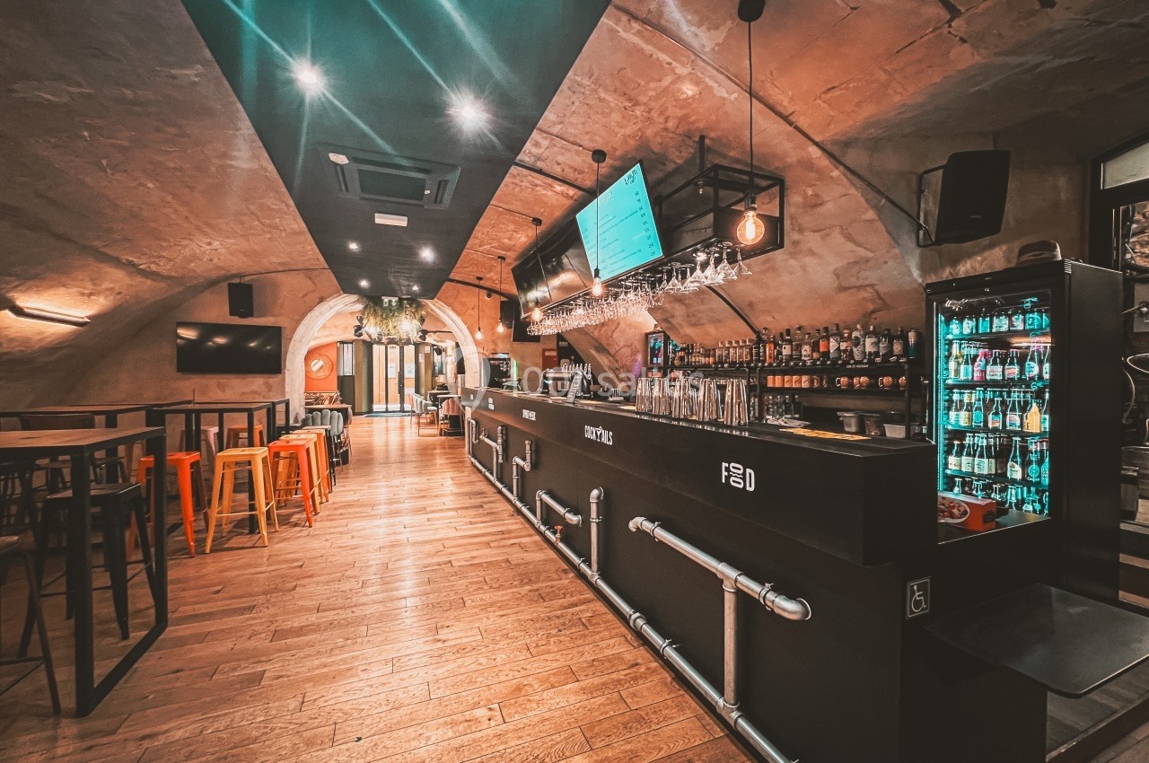 Intérieur d'un bar avec un long comptoir, des tabourets colorés, des étagères de boissons et un éclairage chaleureux.
