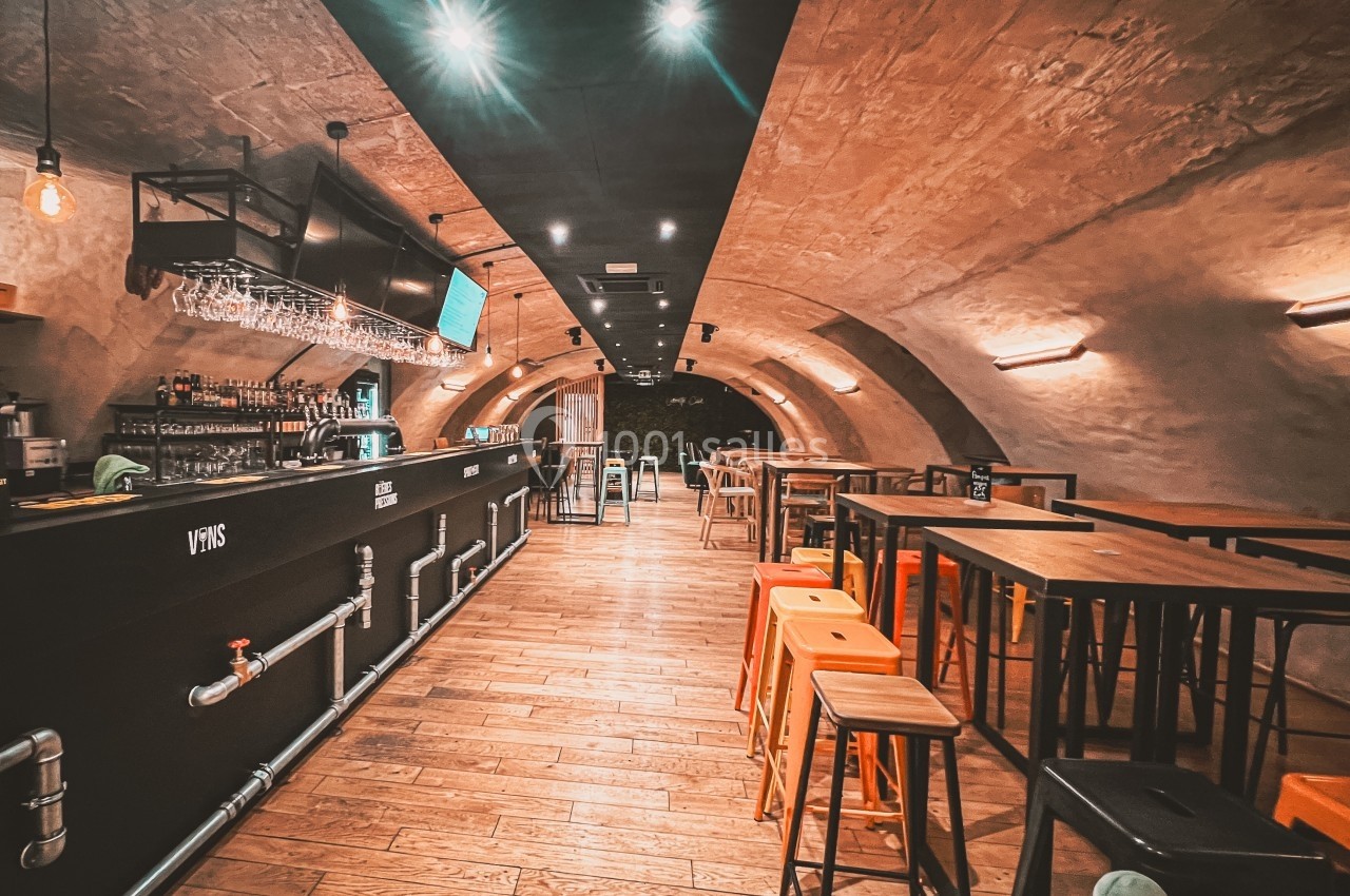 Intérieur d'un bar avec un comptoir long, des tabourets orange et un plafond voûté en pierre éclairé.