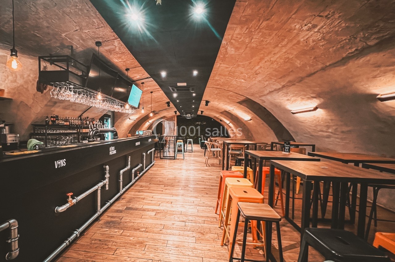 Intérieur d'un bar avec un long comptoir, des tables hautes et des tabourets, dans une salle voûtée en pierre.
