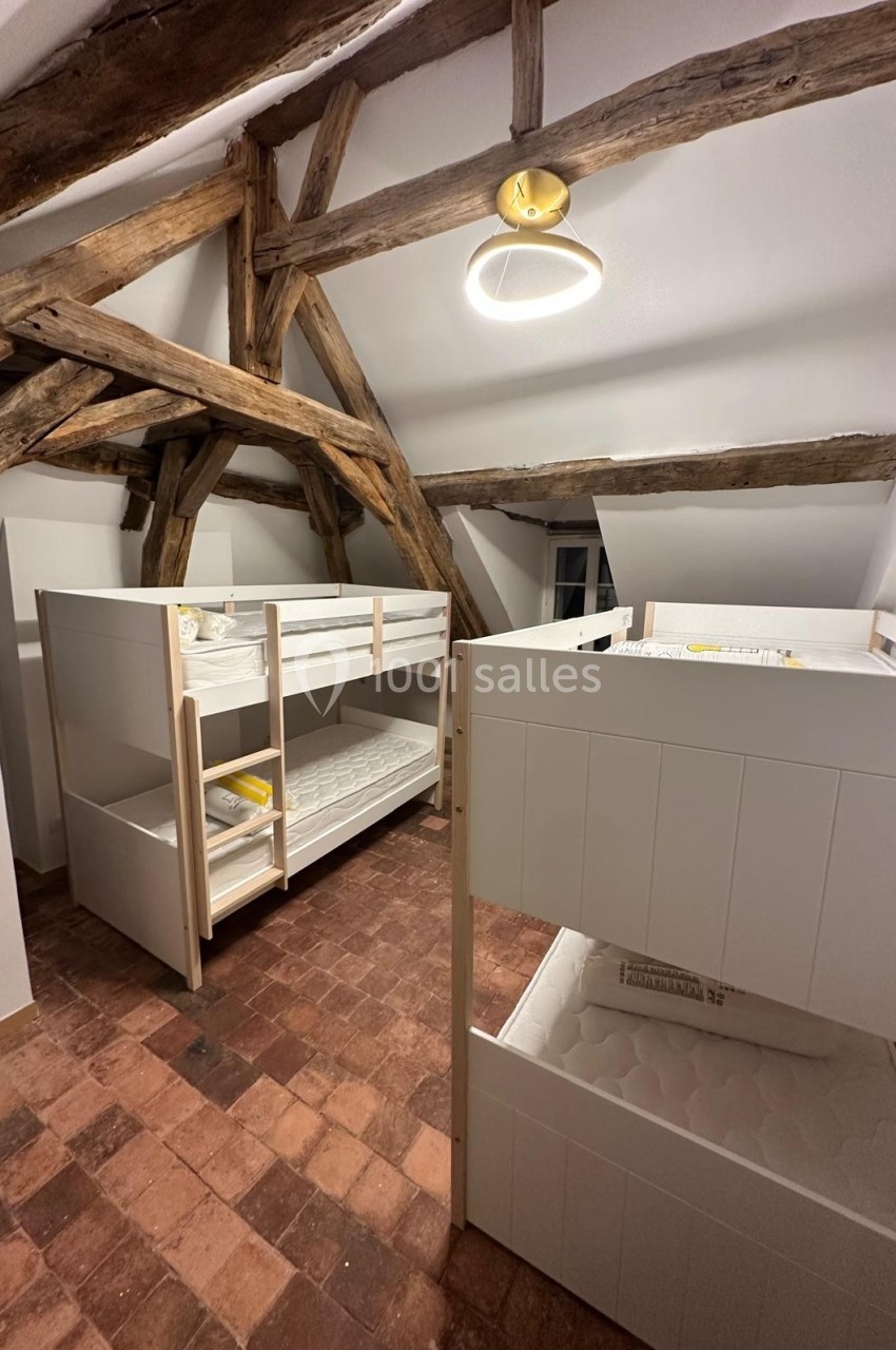 Chambre mansardée avec poutres apparentes, deux lits superposés blancs et sol en tomettes rouges.