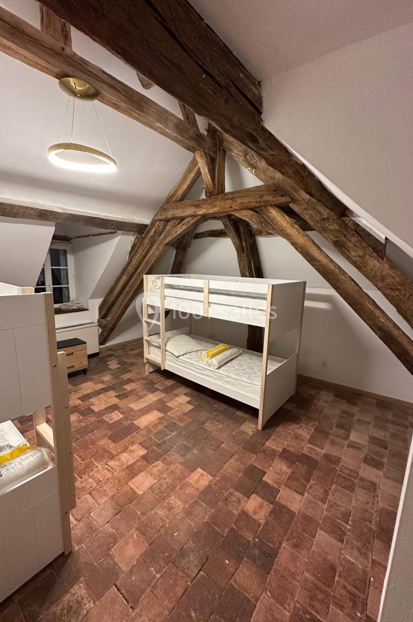 Chambre mansardée avec poutres apparentes, lits superposés en bois clair et sol en carreaux de terre cuite.