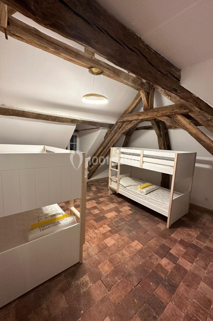 Chambre mansardée avec poutres apparentes, deux lits superposés blancs et sol en tomettes rouges.