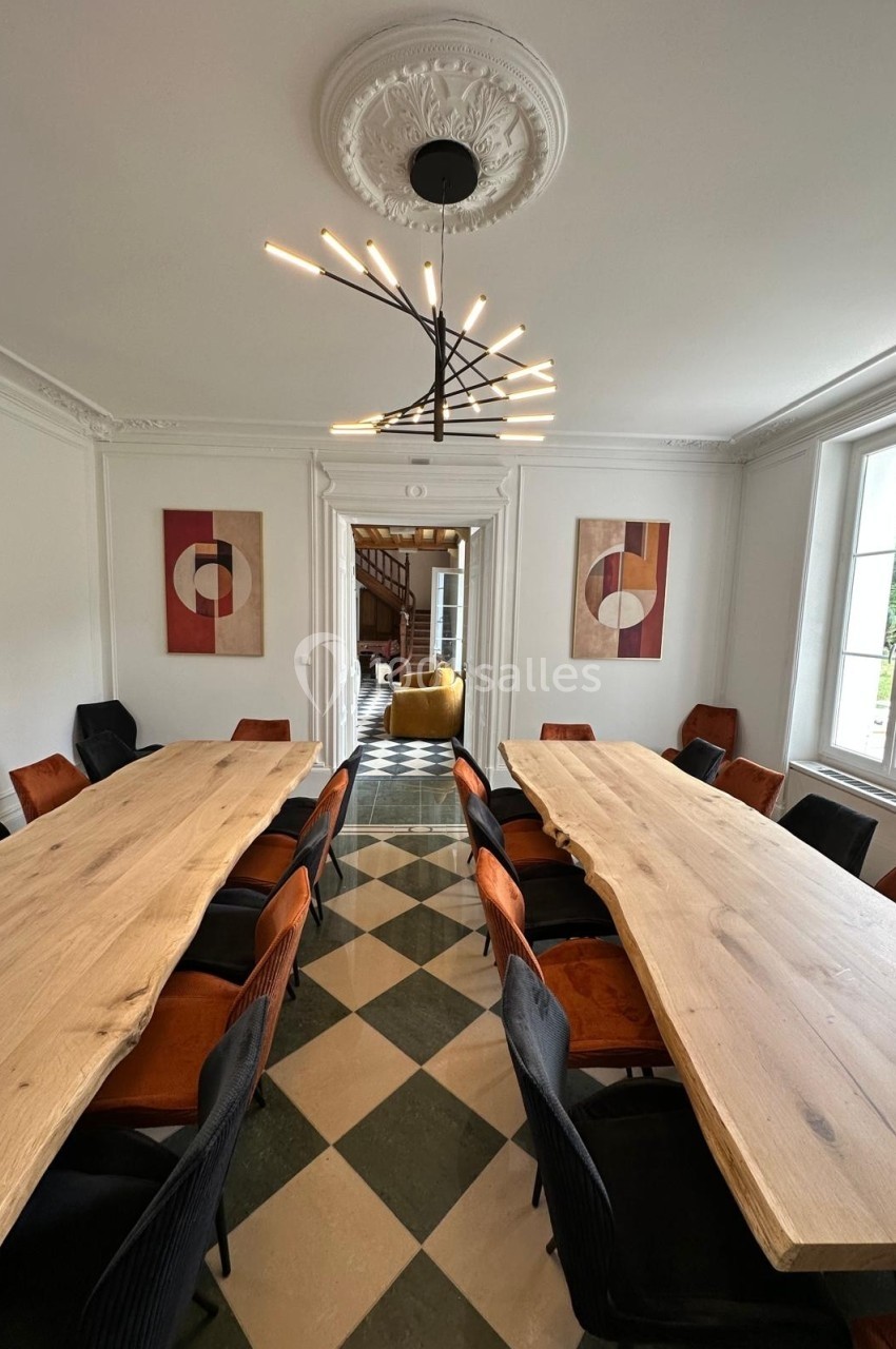 Salle de réunion lumineuse avec deux grandes tables en bois, chaises colorées et lustre moderne, sol à carreaux.