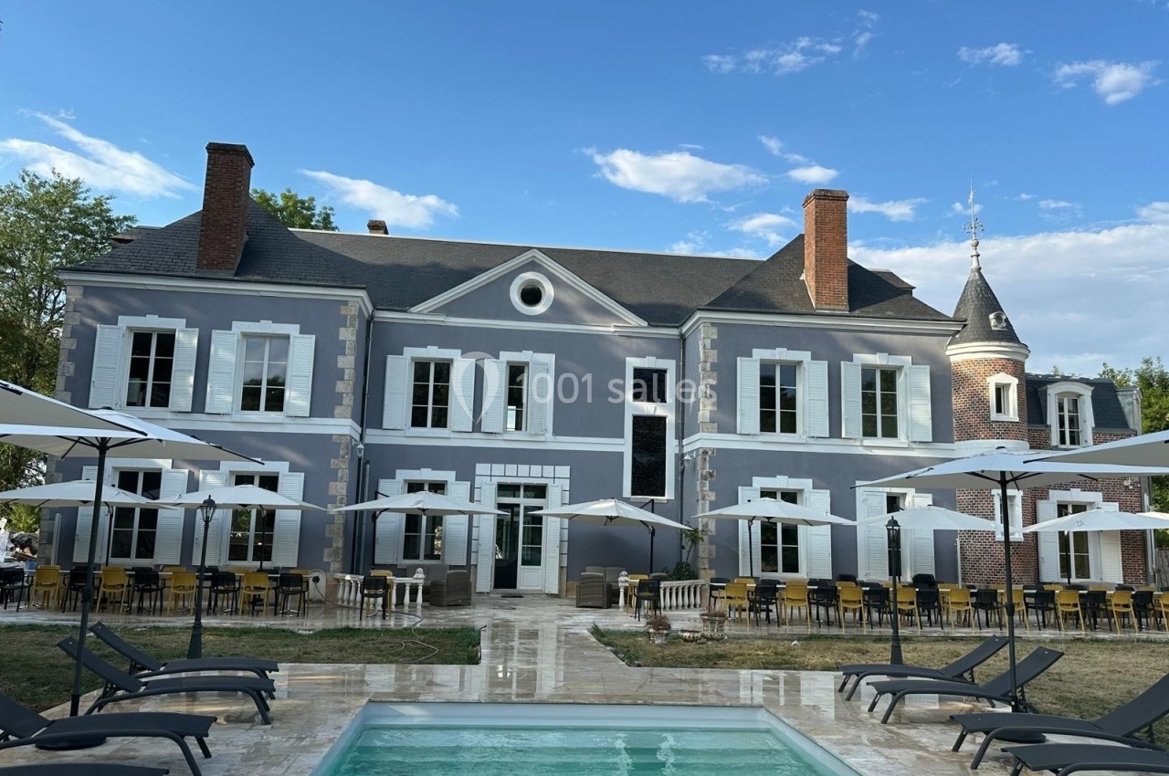 Façade d'un manoir bleu avec terrasse aménagée, parasols et piscine au premier plan, sous un ciel dégagé.