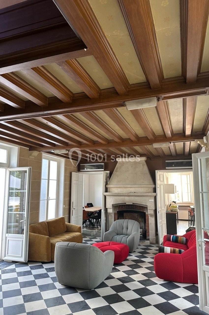 Salle lumineuse avec plafond en bois, cheminée en pierre, mobilier coloré et sol en damier noir et blanc.