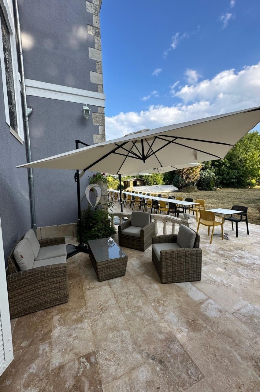 Terrasse avec mobilier en rotin, table, chaises et grand parasol, donnant sur un jardin verdoyant.