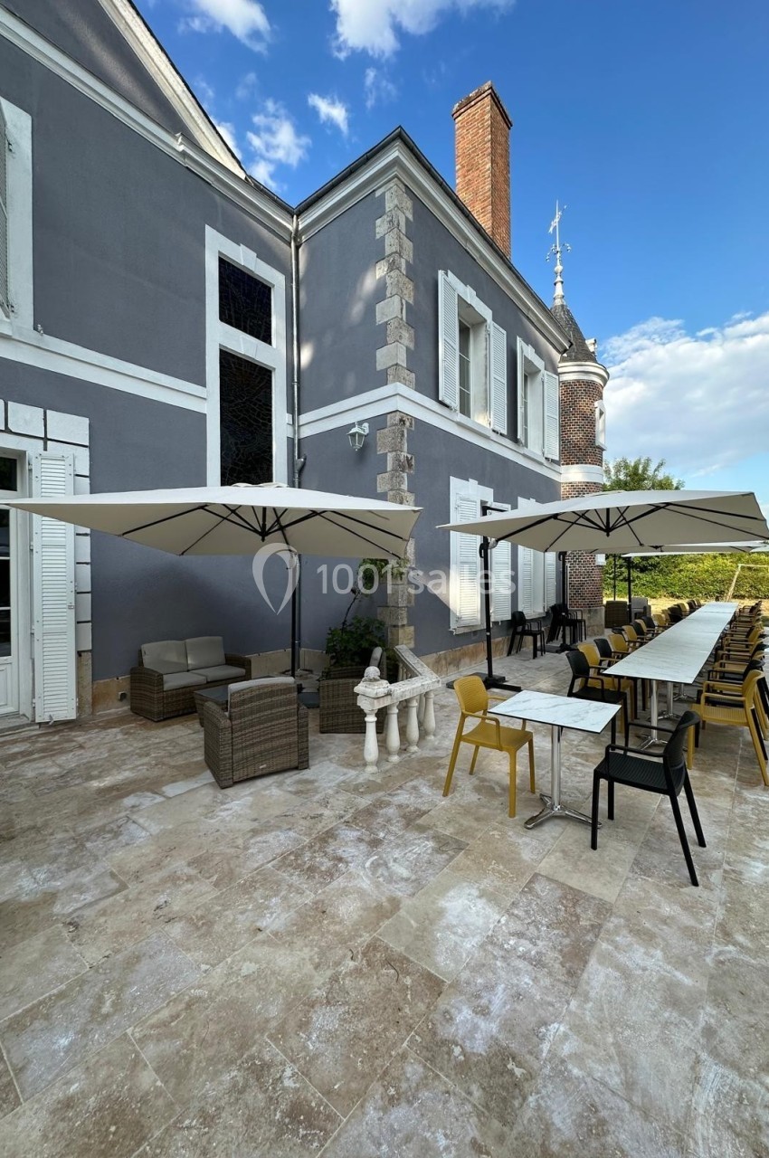 Terrasse en pierre avec tables, chaises, parasols et coin salon, adossée à un bâtiment ancien sous un ciel dégagé.