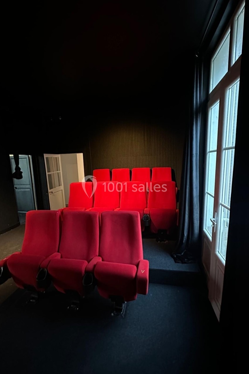 Rangées de fauteuils rouges dans une petite salle sombre avec une porte-fenêtre à droite.