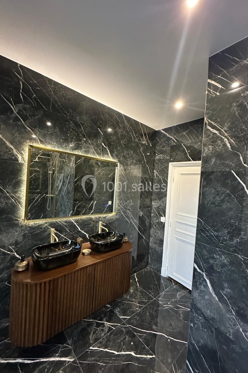 Salle de bain moderne avec murs et sol en marbre noir, double vasque noire sur meuble en bois et miroir éclairé.