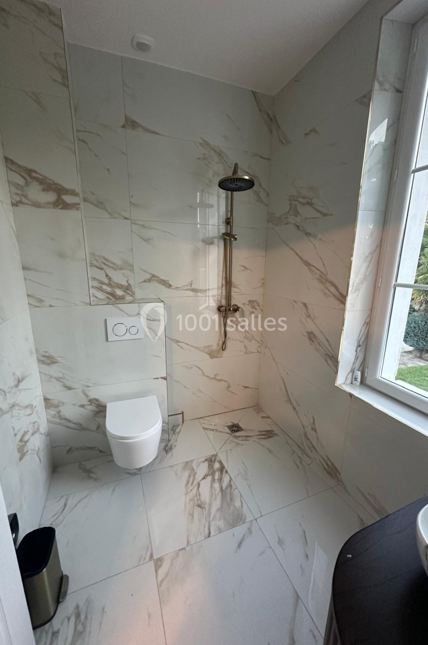 Salle de bain moderne avec toilettes suspendues, douche à l'italienne et carrelage effet marbre près d'une fenêtre.