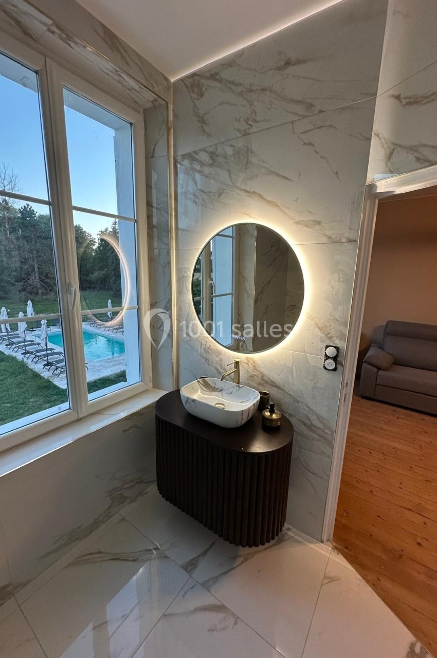 Lavabo moderne avec miroir rond rétroéclairé, devant une fenêtre donnant sur une piscine et un jardin.