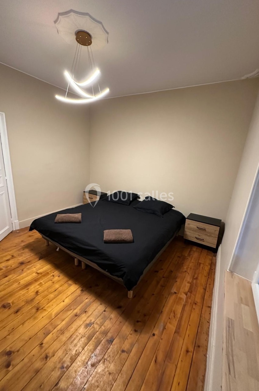 Lit double avec draps noirs, deux serviettes pliées, tables de chevet en bois et sol en parquet dans une chambre sobre.