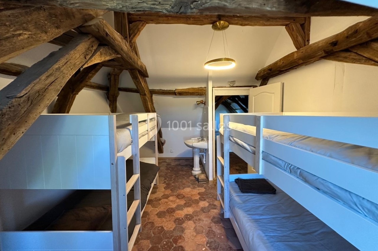Dortoir sous combles avec lits superposés en bois blanc, lavabo central et poutres apparentes au plafond.