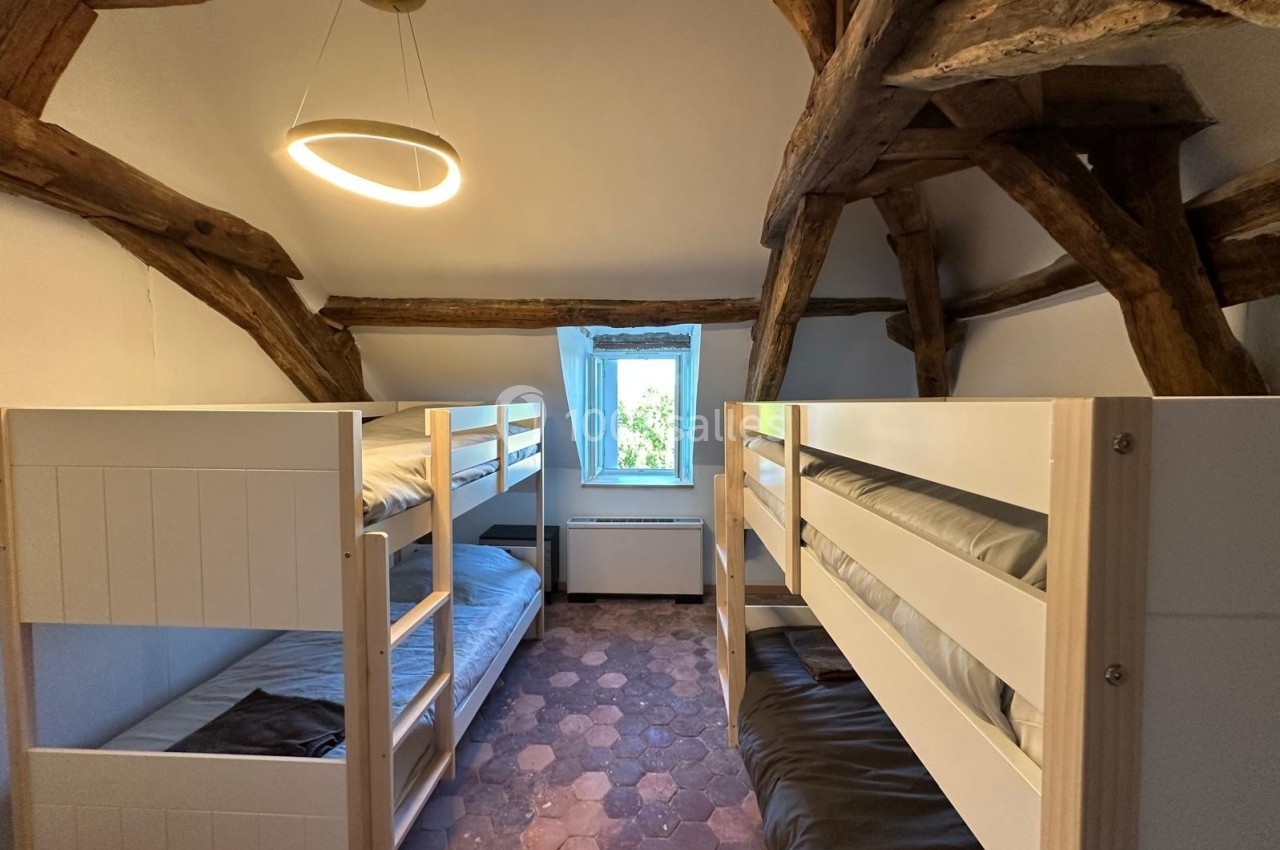 Chambre avec lits superposés en bois, plafond avec poutres apparentes et sol en tomettes, éclairée par une fenêtre.