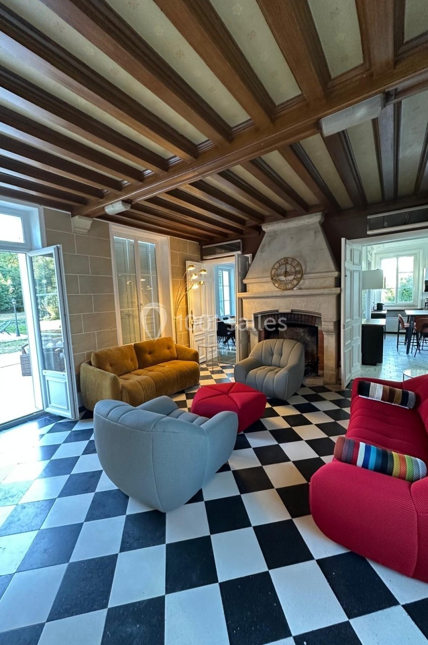 Salon lumineux avec sol en damier, canapés colorés, cheminée en pierre et grandes portes-fenêtres ouvertes sur un jardin.