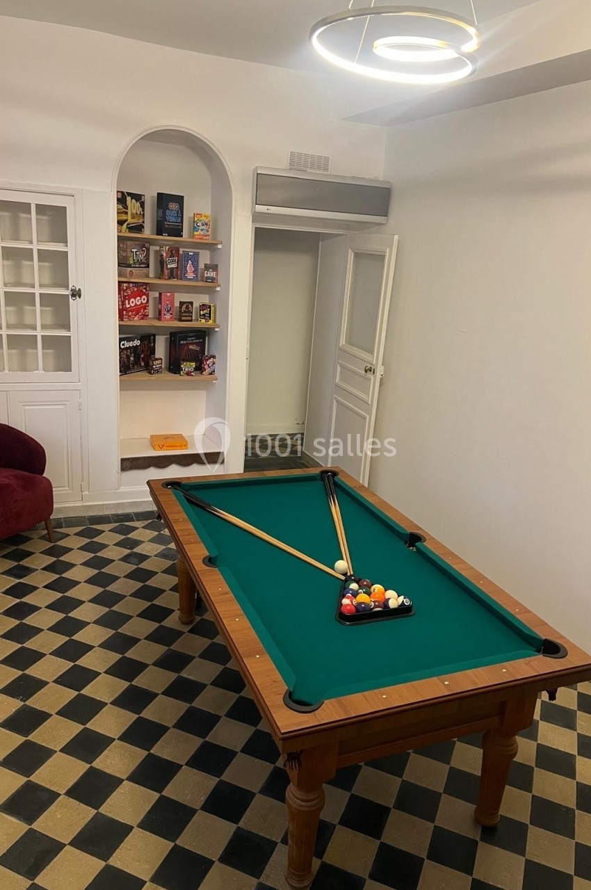 Table de billard avec triangle de boules colorées et deux queues, dans une pièce au sol en damier et étagères murales.