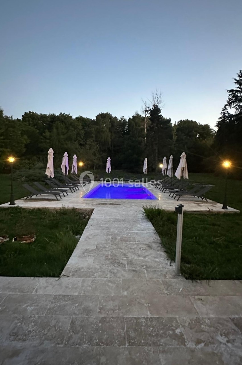Piscine éclairée en soirée, entourée de chaises longues et de parasols, située dans un jardin arboré.