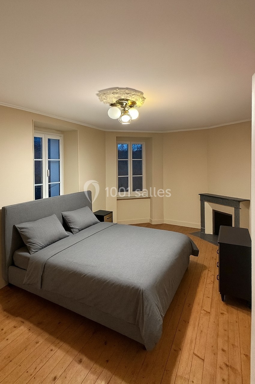 Chambre avec lit double gris, parquet en bois, cheminée, deux fenêtres et un plafonnier lumineux.