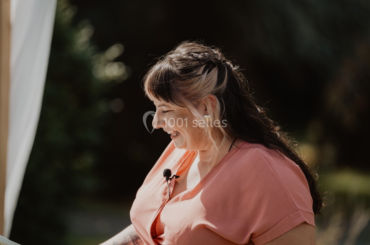 Femme souriante en robe rose, portant un micro-cravate, debout en extérieur avec un fond flou de végétation.