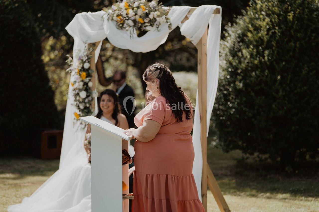 Une femme en robe rose lit un discours devant une mariée souriante, sous une arche décorée de fleurs blanches et jaunes.