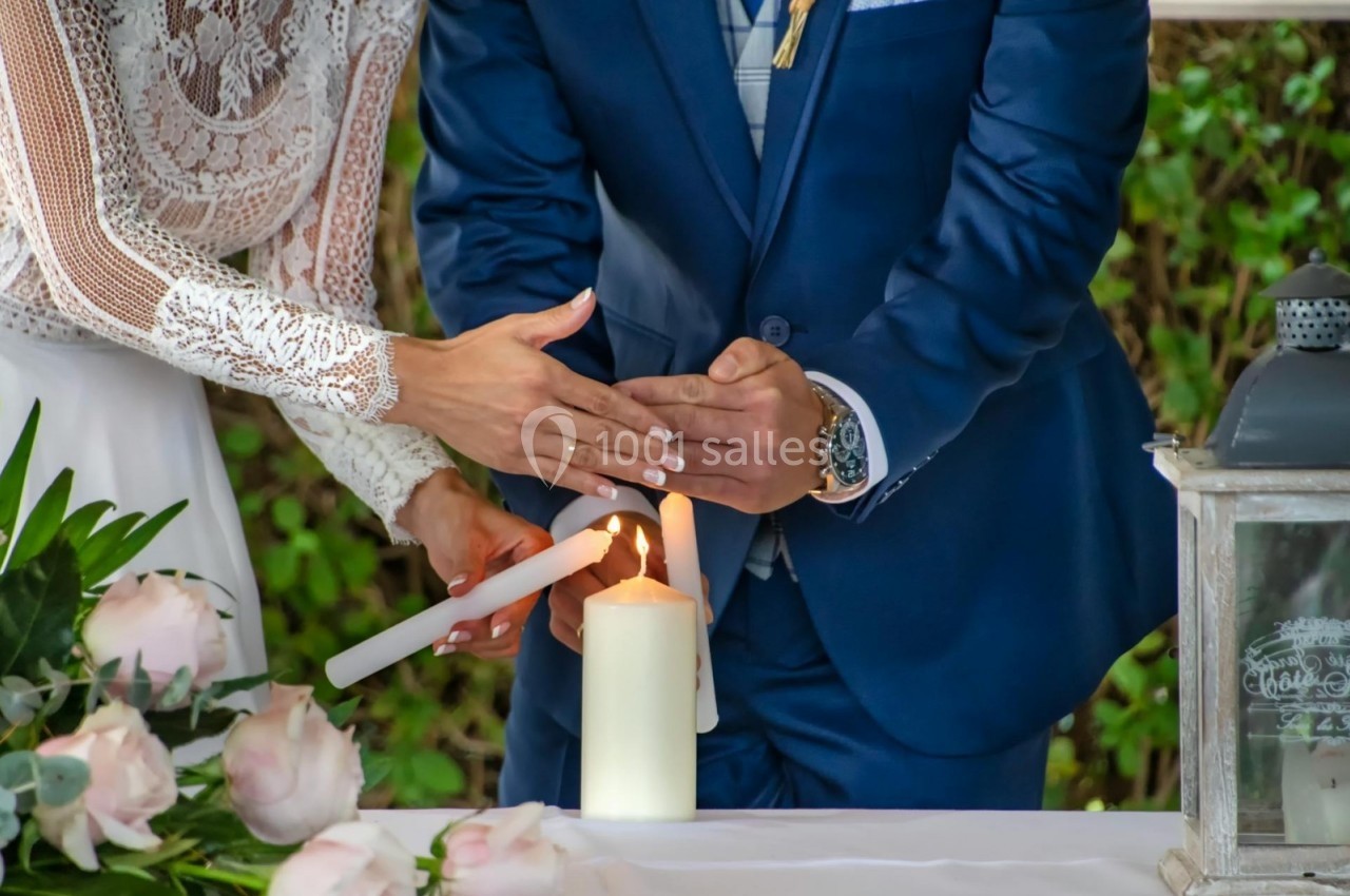 Un couple allume ensemble une bougie blanche lors d'une cérémonie, entouré de fleurs et de verdure.
