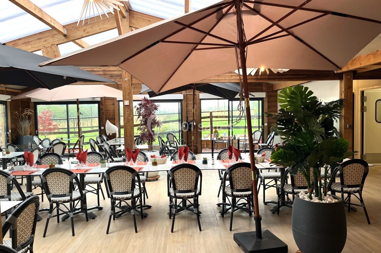 Salle de restaurant lumineuse avec grandes baies vitrées, tables dressées et parasols intérieurs.