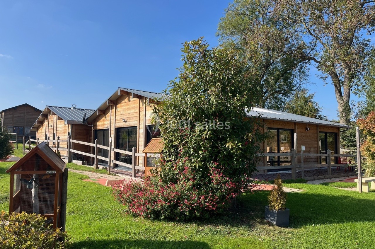 Bâtiments en bois entourés de verdure avec un arbre, des arbustes et un ciel dégagé en arrière-plan.