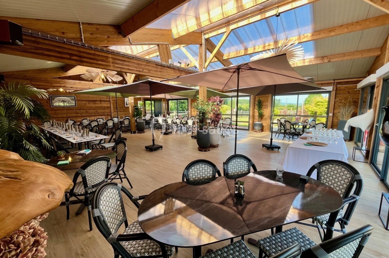 Salle lumineuse avec charpente en bois, grandes baies vitrées, tables dressées et parasols intérieurs.