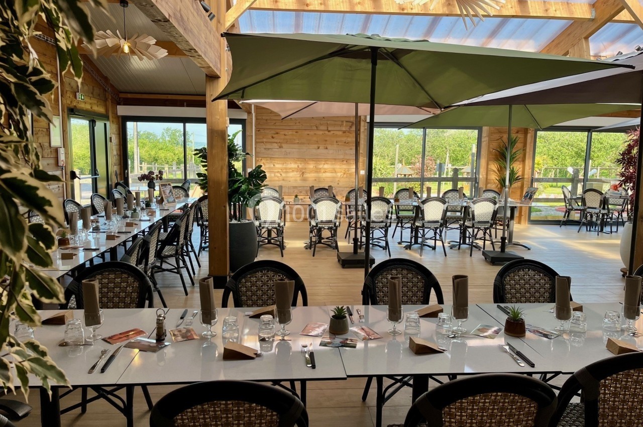Salle lumineuse avec de grandes baies vitrées, tables dressées, chaises en osier et parasols ouverts à l'intérieur.