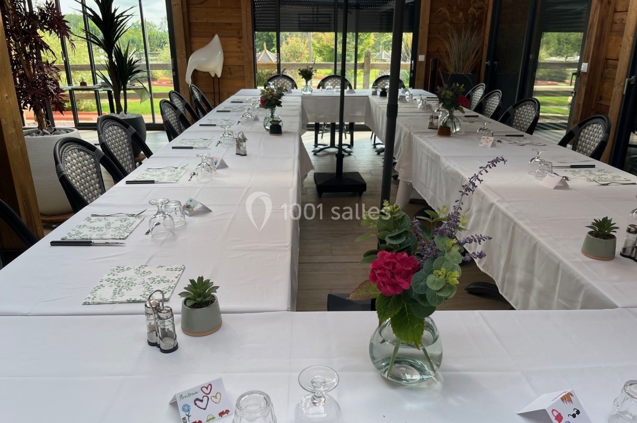 Salle de réunion lumineuse avec tables disposées en U, nappes blanches, décorations florales et vue sur un jardin.