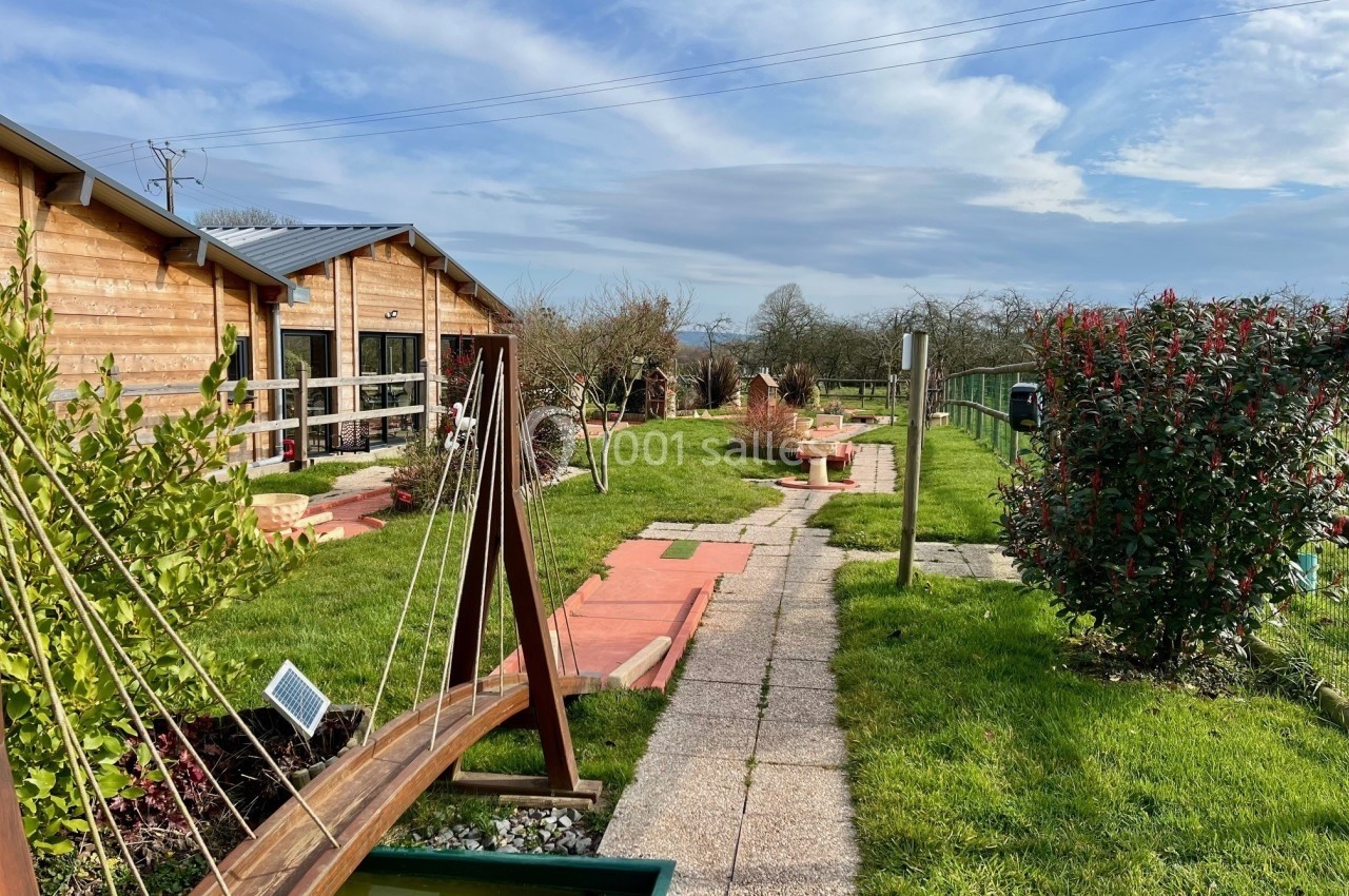 Allée pavée bordée de verdure, avec des structures en bois et un bâtiment en arrière-plan sous un ciel dégagé.