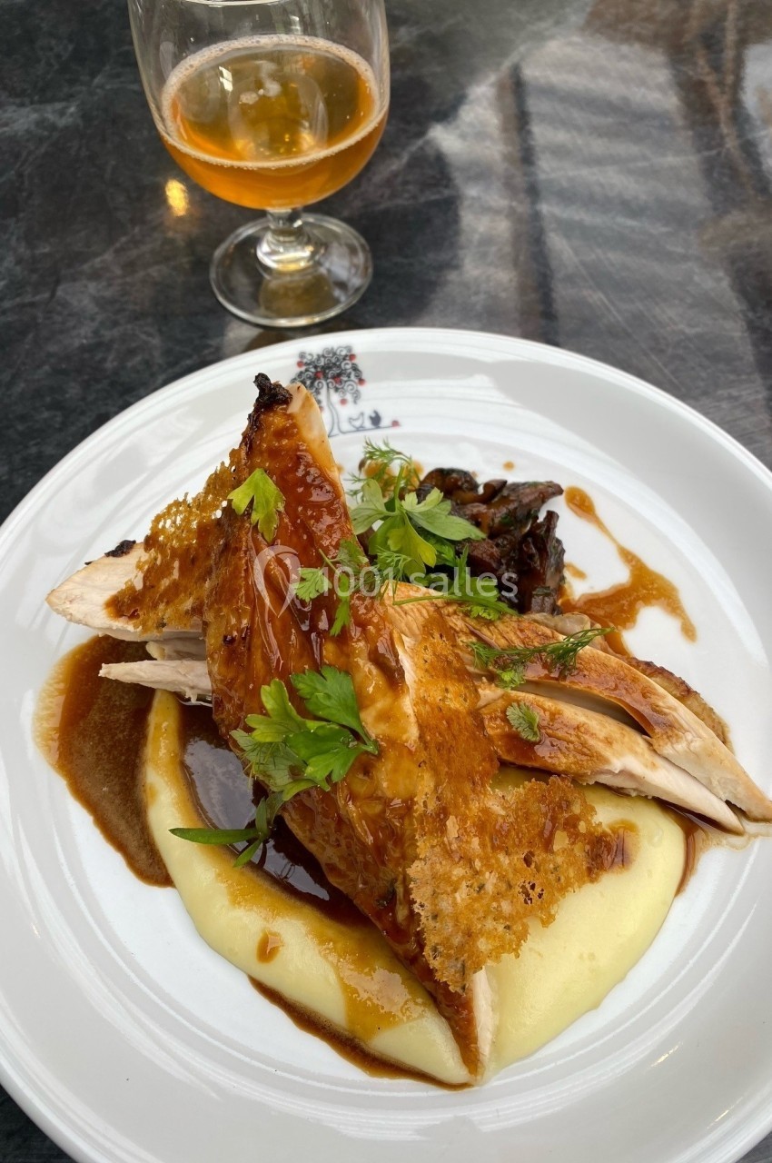 Assiette de volaille rôtie sur purée de pommes de terre, garnie de sauce et herbes, avec un verre de boisson ambrée.