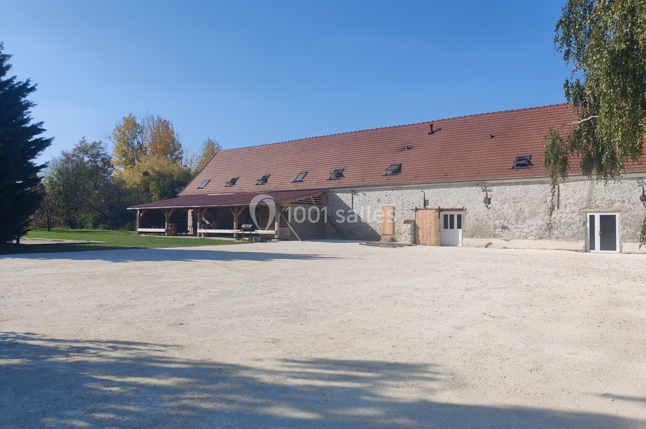 Bâtiment en pierre avec toit en tuiles rouges, grande cour dégagée et arbres en arrière-plan sous un ciel bleu.