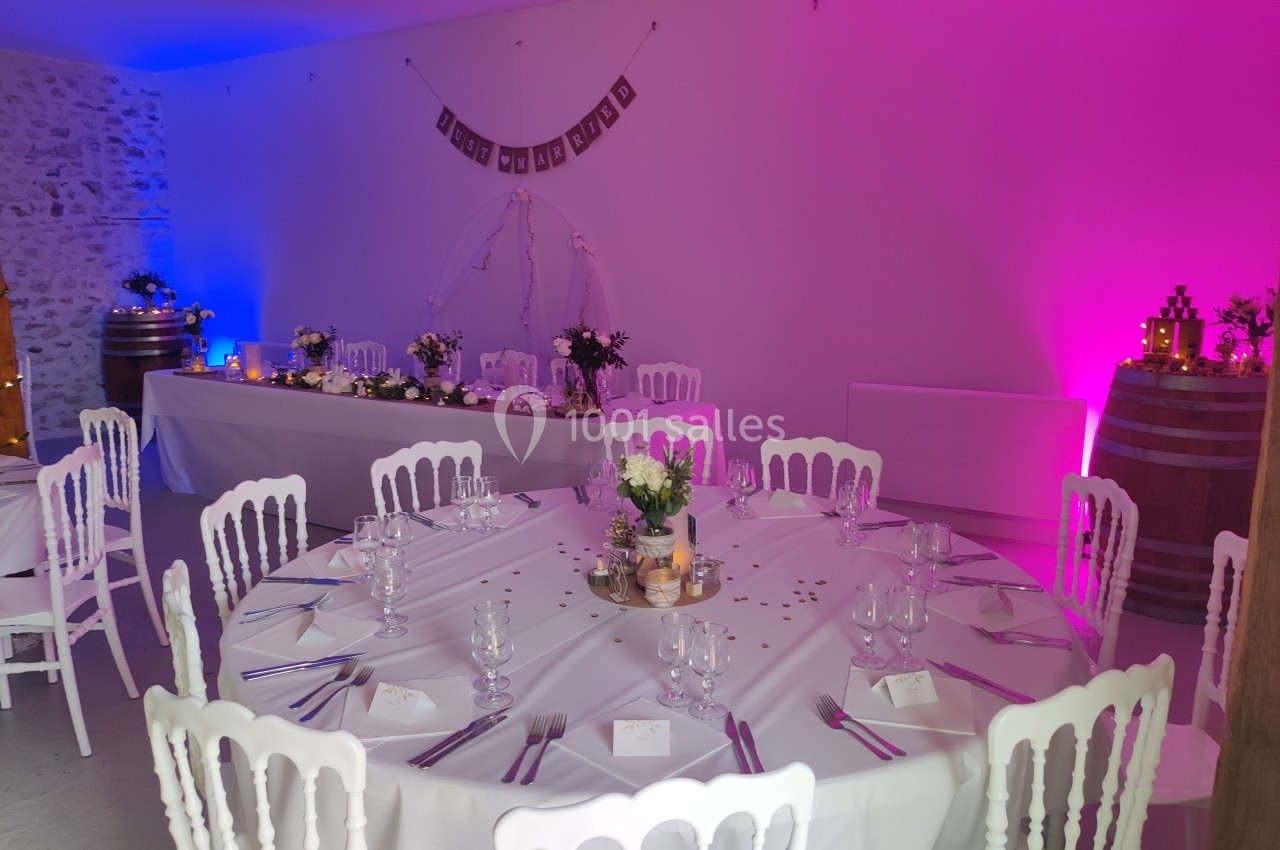Salle décorée pour un mariage avec tables rondes dressées, guirlande murale et éclairage rose et bleu.
